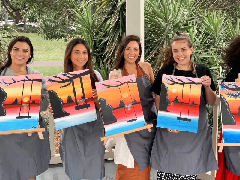 Four women holding up their artworks at outdoor paint and sip class